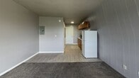 Located at 4213 Lake Land Blvd in Mattoon, IL, this compact single-floor unit offers 270 square feet of functional living space, comprising one bedroom and one bathroom. The open layout seamlessly connects the main living area to the kitchen, maximizing the use of every square foot. The living and bedroom area spans approximately 230 square feet and features gray carpet flooring, neutral gray-painted walls, and a window with blinds that provides natural light and a pleasant outdoor view of the surrounding greenery. The kitchen is efficiently designed with warm wood cabinetry, a white refrigerator, and a sink, all set on durable wood-look vinyl flooring that flows through the entry corridor toward the front door. An electrical panel is visibly integrated into the main living space wall. The bathroom is accessed through a paneled door and includes a white vanity with cabinet storage, a wall-mounted mirror, sconce lighting, a toilet, and tile-accented walls, adding a classic touch to the space. The unit's straightforward single-level design and neutral color palette create a clean, functional environment well-suited for individuals seeking an efficient and low-maintenance living space in Mattoon, Illinois.
