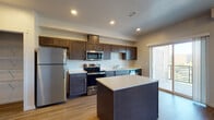 Located at 1608 East Riverton Avenue in Spokane, Washington, this well-designed single-floor unit offers a thoughtful layout with modern finishes throughout. The kitchen serves as a central highlight, showcasing rich dark cabinetry, light quartz countertops, a full suite of stainless steel appliances including a refrigerator, range, microwave, and dishwasher, and a center island that provides additional workspace and storage. Light wood-style flooring runs throughout the main living areas, lending warmth and continuity to the space. The living area connects seamlessly to a balcony through a sliding glass door, offering pleasant outdoor views of surrounding trees. The bedrooms are carpeted for comfort, with one featuring a walk-in closet with built-in shelving and another offering a standard closet with direct access to an adjoining bathroom. The bathrooms are finished with dark vanity cabinetry, quartz countertops, and wood-style flooring. One bathroom includes a full tub-shower combination with a high window that allows natural light, while another features a walk-in closet-style linen storage area. Corner windows in one of the bedrooms frame expansive tree-lined views, filling the room with abundant natural light. The unit presents a clean, contemporary aesthetic throughout.