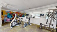 Located at 540 S Carroll Rd in Villa Rica, Georgia, this well-appointed fitness center offers a comprehensive exercise environment within a single, efficiently organized room. The space is finished with classic architectural details, including crown molding, white wainscoting panels along the lower walls, and recessed lighting that creates a bright, welcoming atmosphere. A ceiling fan overhead aids in air circulation during workouts.

Large multi-pane windows dominate one wall, flooding the room with natural light and providing direct views of an outdoor pool and surrounding tree line — an inspiring backdrop for any workout. A wall-mounted flat-screen television is positioned near the windows for added entertainment.

The equipment layout is well-considered, featuring treadmills, elliptical trainers, upright and recumbent stationary bikes, a cable strength machine, and a rowing/bench station. A dedicated stretching and flexibility area includes exercise balls and additional accessories. Floor-to-ceiling mirrors line one wall, visually expanding the space and allowing users to monitor their form.

Adding a bold artistic touch, a large, colorful abstract mural anchors one end of the room, energizing the overall ambiance. Durable, patterned modular carpet tiles cover the floor throughout, providing comfort and traction. This fitness center is a functional and visually engaging space suited for a full range of workout routines.