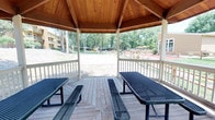 This outdoor recreational area centers around a charming gazebo pavilion constructed with warm, natural wood-paneled vaulted ceilings supported by white-painted posts. The open-air structure features a classic octagonal design and sits atop a wood deck, furnished with multiple metal picnic tables and benches ideal for group gatherings. White-painted railings frame the perimeter, offering unobstructed views of the surrounding grounds from every angle.

Beyond the gazebo, the complex offers an impressive array of outdoor amenities. A well-maintained sand volleyball court sits amid mature, leafy trees, providing natural shade and a lush backdrop. A full-sized tennis court with blue surfacing doubles as a basketball court, complete with a hoop and net, enclosed by a chain-link fence. Additional recreational features visible from the gazebo include a children's playground and shaded picnic areas with umbrellas.

Concrete pathways wind through the landscaped grounds, connecting the various activity areas. Mature trees throughout the property provide ample shade and greenery. The multi-building residential complex surrounds the recreational space, creating a cohesive community environment. The overall ambiance is active, open, and well-suited for outdoor recreation and social interaction.