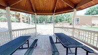 This outdoor recreational area centers around a charming gazebo pavilion constructed with warm, natural wood-paneled vaulted ceilings supported by white-painted posts. The open-air structure features a classic octagonal design and sits atop a wood deck, furnished with multiple metal picnic tables and benches ideal for group gatherings. White-painted railings frame the perimeter, offering unobstructed views of the surrounding grounds from every angle.

Beyond the gazebo, the complex offers an impressive array of outdoor amenities. A well-maintained sand volleyball court sits amid mature, leafy trees, providing natural shade and a lush backdrop. A full-sized tennis court with blue surfacing doubles as a basketball court, complete with a hoop and net, enclosed by a chain-link fence. Additional recreational features visible from the gazebo include a children's playground and shaded picnic areas with umbrellas.

Concrete pathways wind through the landscaped grounds, connecting the various activity areas. Mature trees throughout the property provide ample shade and greenery. The multi-building residential complex surrounds the recreational space, creating a cohesive community environment. The overall ambiance is active, open, and well-suited for outdoor recreation and social interaction.