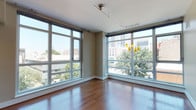 This contemporary single-floor space is defined by its striking floor-to-ceiling windows that wrap around the living area, flooding the interior with natural light and framing dynamic urban and treetop views of the surrounding cityscape. Rich hardwood flooring flows throughout the main living areas, lending warmth and continuity to the open layout.

The kitchen is well-appointed, featuring dark espresso cabinetry extending to the ceiling for generous storage, granite countertops, a white tile backsplash, and stainless steel appliances including a refrigerator, range, and microwave. A peninsula provides additional counter space and casual seating potential, while a window alongside the kitchen adds further natural light. Track lighting and recessed ceiling fixtures illuminate both the living and kitchen areas effectively.

The bedroom offers plush carpeting and a walk-in closet with built-in shelving for organized storage. Two bathrooms serve the space: one features a sleek glass-enclosed shower with floor-to-ceiling wood-tone tile, a built-in niche, and a corner bench, while the second includes a soaking tub with matching tile surround, a granite-topped vanity with dark cabinetry, and warm tile flooring. Together, these elements create a polished, urban aesthetic throughout.