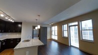 This single-floor space showcases a thoughtfully designed open-concept layout that seamlessly connects the kitchen and living areas. The kitchen features a granite-topped island with an undermount sink and stylish pendant lighting hanging above, providing both functional workspace and a visual focal point. Dark hardwood flooring flows throughout, creating a rich contrast against the neutral-toned walls and white ceiling.

The living area benefits from abundant natural light streaming through multiple large windows, as well as what appears to be a glass door providing access to an outdoor space, such as a balcony or patio. Track lighting in the living area supplements the natural light and adds a contemporary touch.

A gray interior door leads to an adjacent area, where a stacked washer and dryer unit is visible, indicating convenient in-unit laundry. The overall layout feels open and airy, with a raised ceiling detail in the kitchen zone adding architectural interest. The combination of modern finishes, warm flooring, and well-placed lighting gives the space a clean, contemporary ambiance well-suited for everyday living.