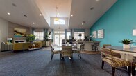 Located at 3900 75th St W in Bradenton, Florida, this single-story community clubhouse and leasing center is designed with a coastal-inspired aesthetic that immediately sets an inviting tone. Bold teal accent walls contrast with crisp white ceilings and neutral gray carpeting throughout, creating a visually dynamic yet cohesive atmosphere.

The main reception area greets visitors with soaring vaulted ceilings, an elegant pendant chandelier, and a large front desk flanked by arched glass entry doors that flood the space with natural light. Adjacent to the reception desk, glass-partitioned private offices provide a professional setting for one-on-one consultations, each thoughtfully furnished and decorated with coastal artwork.

The open lounge and gathering area is generously proportioned, featuring multiple seating arrangements with colorful, patterned upholstery, round dining tables, and a built-in bar-height counter — ideal for community events or casual interaction. A cozy fireplace lounge with a textured accent wall and comfortable seating offers a quieter retreat within the space.

A secondary lounge area, decorated with vibrant yellow and teal furnishings and beach-themed artwork, adds a relaxed, resort-like feel. French doors at the rear open directly to an outdoor pool area, seamlessly connecting indoor and outdoor living. Natural greenery placed throughout enhances the fresh, tropical ambiance of this well-appointed facility.