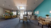 Located at 3900 75th St W in Bradenton, Florida, this single-story community clubhouse and leasing center is designed with a coastal-inspired aesthetic that immediately sets an inviting tone. Bold teal accent walls contrast with crisp white ceilings and neutral gray carpeting throughout, creating a visually dynamic yet cohesive atmosphere.

The main reception area greets visitors with soaring vaulted ceilings, an elegant pendant chandelier, and a large front desk flanked by arched glass entry doors that flood the space with natural light. Adjacent to the reception desk, glass-partitioned private offices provide a professional setting for one-on-one consultations, each thoughtfully furnished and decorated with coastal artwork.

The open lounge and gathering area is generously proportioned, featuring multiple seating arrangements with colorful, patterned upholstery, round dining tables, and a built-in bar-height counter — ideal for community events or casual interaction. A cozy fireplace lounge with a textured accent wall and comfortable seating offers a quieter retreat within the space.

A secondary lounge area, decorated with vibrant yellow and teal furnishings and beach-themed artwork, adds a relaxed, resort-like feel. French doors at the rear open directly to an outdoor pool area, seamlessly connecting indoor and outdoor living. Natural greenery placed throughout enhances the fresh, tropical ambiance of this well-appointed facility.