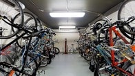 This single-floor indoor bicycle storage room is purpose-built to accommodate a large number of bicycles in an organized and efficient manner. The room is lined on both sides with robust bike racks, which hold a diverse array of bicycles — from road bikes to mountain bikes — in a vertical or angled orientation to maximize the use of available floor space. A central aisle runs the length of the room, providing clear and easy access to each stored bicycle. The space is well-lit by overhead fluorescent lighting fixtures mounted to the ceiling, ensuring visibility throughout the entire area at all times. The floors are finished with a smooth, durable surface suited to the demands of a high-traffic storage environment. A standout feature of this space is a dedicated bike repair station located toward the back of the room, equipped with a floor pump and a repair stand, allowing cyclists to perform maintenance and adjustments with ease. Exposed utility conduit runs along the ceiling, reflecting the functional, no-frills design philosophy of the space. The neutral-toned walls contribute to a clean, utilitarian aesthetic. Overall, this bicycle storage room is a well-organized, practical facility designed to serve the needs of an active cycling community.