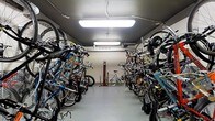 This single-floor indoor bicycle storage room is purpose-built to accommodate a large number of bicycles in an organized and efficient manner. The room is lined on both sides with robust bike racks, which hold a diverse array of bicycles — from road bikes to mountain bikes — in a vertical or angled orientation to maximize the use of available floor space. A central aisle runs the length of the room, providing clear and easy access to each stored bicycle. The space is well-lit by overhead fluorescent lighting fixtures mounted to the ceiling, ensuring visibility throughout the entire area at all times. The floors are finished with a smooth, durable surface suited to the demands of a high-traffic storage environment. A standout feature of this space is a dedicated bike repair station located toward the back of the room, equipped with a floor pump and a repair stand, allowing cyclists to perform maintenance and adjustments with ease. Exposed utility conduit runs along the ceiling, reflecting the functional, no-frills design philosophy of the space. The neutral-toned walls contribute to a clean, utilitarian aesthetic. Overall, this bicycle storage room is a well-organized, practical facility designed to serve the needs of an active cycling community.