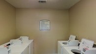 The Laundry Center is a dedicated, single-floor communal laundry facility designed to serve residents with practical, everyday convenience. The room features a straightforward layout with white top-loading washing machines and dryers positioned along both side walls, maximizing floor space and allowing comfortable access to each unit. The walls are finished in a neutral beige tone, contributing to a clean and utilitarian atmosphere. A textured ceiling with a centrally placed ventilation duct ensures adequate airflow throughout the space. Prominently displayed on the back wall is a "Laundry Center" rules sign, outlining usage guidelines and emergency contact information, reflecting an organized and well-managed facility. The overall ambiance is functional and no-frills, prioritizing efficiency and ease of use. The space is well-suited for its intended purpose, offering residents a reliable and orderly environment for laundry needs.