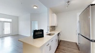 This single-floor residence offers a clean, contemporary aesthetic highlighted by an open-concept layout that seamlessly connects the kitchen and living areas. The kitchen is a standout feature, showcasing bright white cabinetry with brushed nickel hardware, a light quartz countertop that extends into a peninsula for additional prep or seating space, and a stainless steel undermount sink with a modern pull-down faucet. A tile backsplash adds a subtle design element behind the counter, while a stainless steel dishwasher provides everyday convenience. Recessed track lighting above the kitchen ensures ample illumination, complementing the flush-mount ceiling light in the adjoining space. The flooring throughout features warm-toned hardwood-style planks that contrast beautifully with the crisp white walls, creating a balanced and inviting ambiance. The open sightlines into the adjacent room suggest a fluid, connected floor plan ideal for both everyday living and entertaining. The overall design embraces a minimalist, modern character with neutral tones, quality materials, and thoughtful details that contribute to a bright, welcoming environment.