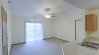 This single-floor space offers a clean, functional layout designed for comfortable everyday living. The main living area immediately draws attention with its vaulted ceiling, which lends a sense of openness and volume to the room. A ceiling fan with an integrated light fixture is centrally mounted, providing both air circulation and ambient lighting. Plush wall-to-wall carpeting in a neutral tone covers the floor, adding warmth and comfort underfoot. A large sliding glass door dominates one wall, dressed with vertical blinds that allow for adjustable light control. The door floods the space with natural light and provides direct access to an outdoor area, creating a seamless connection between interior and exterior. The walls are finished in a crisp, neutral white that enhances the brightness throughout. Warm wood-toned baseboards frame the perimeter, adding a subtle architectural detail. A doorway is visible on the adjacent wall, suggesting additional rooms beyond the main living area. The overall ambiance is light, airy, and move-in ready, offering a versatile canvas well-suited for a variety of furnishing styles.