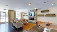 This single-floor space is designed as a contemporary lounge or common area, blending modern aesthetics with comfortable, functional furnishings. The focal point of the room is a floor-to-ceiling accent wall clad in chevron-patterned wood-look tile, which houses a sleek linear fireplace and a wall-mounted flat-screen television, creating a striking visual centerpiece.

The seating area is anchored by a neutral-toned sectional sofa paired with a round glass coffee table and a warm amber shag area rug, adding texture and color contrast to the polished concrete flooring. Compact orange upholstered cube ottomans introduce vibrant accent tones throughout the space. A casual dining area with a round wood table and modern white chairs is positioned nearby, offering additional functionality.

Large windows along one wall allow ample natural light to fill the room, complemented by recessed ceiling lighting. Decorative wall-mounted planters featuring ornamental grasses are arranged throughout, adding a fresh, organic element to the interior. Neutral green drapery softens the windows and contributes to the space's calm, welcoming ambiance.