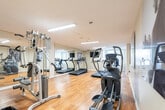 Located at 57 Queen St N in Kitchener, Ontario, this fitness centre is a well-equipped, single-floor facility designed to support a range of workout routines. The space is accessed through a tiled entryway corridor, which transitions into the main exercise area featuring warm hardwood flooring throughout. The room is illuminated by fluorescent overhead lighting and benefits from natural light courtesy of windows fitted with vertical blinds along one wall.

The cardio section is well-appointed, offering multiple treadmills, an elliptical trainer, and a stationary bike, all arranged to allow comfortable use. A full-length wall mirror spans one side of the room, visually expanding the space and allowing users to monitor their form. Strength training needs are addressed by a multi-function cable machine and additional resistance equipment. A wall-mounted flat-screen television provides entertainment during workouts.

Motivational fitness photography is displayed as large-format wall art, adding energy and visual interest to the space. Practical amenities include a hand sanitizer dispenser near the entrance and waste bins positioned throughout. The neutral, light-coloured walls and clean layout contribute to a functional and focused workout environment, making this fitness centre a convenient and well-maintained amenity.