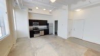 This single-floor unit showcases a distinctive loft-inspired aesthetic defined by dramatically high ceilings with exposed white-painted beams, ductwork, and decorative tin ceiling details. Polished concrete floors run throughout the open-plan living area, reinforcing the industrial character of the space. Large multi-pane windows along one wall admit generous natural light and provide views of the surrounding streetscape, while painted brick walls add textural depth to the interior.

The kitchen is thoughtfully outfitted with dark espresso cabinetry extending to the upper walls, granite-style countertops, a stainless steel sink, and a full suite of stainless steel appliances including a refrigerator, range, built-in microwave, and dishwasher. The kitchen integrates seamlessly into the open living area, making it well-suited for entertaining.

A separate room with its own window offers flexibility as a bedroom or private workspace, continuing the exposed ceiling aesthetic. The bathroom is clean and functional, featuring a full-size soaking tub with an overhead shower, white tile surround, and chrome fixtures. The overall ambiance is open, airy, and urban, with architectural character that distinguishes it from conventional residential spaces.
