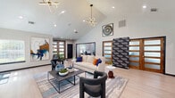 Located on Audelia Road in Dallas, Texas, this thoughtfully designed single-floor space makes a striking first impression from the moment you enter. The welcoming entrance flows seamlessly into an expansive living room anchored by soaring vaulted ceilings that amplify the sense of openness and airiness throughout. A standout floor-to-ceiling geometric-patterned tile fireplace serves as the dramatic focal point of the living area, complemented by a wall-mounted TV and an eye-catching modern pendant light fixture overhead. Light hardwood flooring runs throughout, adding warmth and continuity to the space. Large windows invite abundant natural light, enhancing the bright, airy atmosphere. Contemporary abstract artwork adorns the walls, adding vibrant pops of color against the neutral palette. Warm wood-framed interior doors with frosted glass panels add an elegant architectural detail, providing both privacy and visual interest. The dining area transitions naturally from the living space and features a compact workspace station with dark accent walls, pendant lighting, and built-in computer terminals — a practical and stylish addition. A round dining table with modern blue chairs completes this area, offering a functional yet refined setting. Together, the spaces create a cohesive, design-forward environment ideal for gathering and productivity.