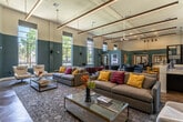 This expansive single-floor space is designed as a multifunctional community hub, characterized by soaring ceilings, rich herringbone-patterned hardwood floors, and a striking two-tone color scheme of teal and cream that runs throughout. Linear pendant lighting suspended from the ceiling enhances the airy, open atmosphere.

The main lounge area is generously proportioned, featuring multiple distinct seating zones furnished with contemporary sofas, accent chairs in blue, red, and neutral tones, and glass-topped coffee tables. Large windows along the perimeter flood the space with natural light and frame views of surrounding greenery.

A stylish bar counter with dark tile cladding and mustard-yellow barstools anchors one side of the space, complemented by open shelving displaying decorative accents. A shuffleboard table and a glass-enclosed fitness room with red velvet curtain accents add recreational elements.

A dedicated work zone includes a dark executive desk with red accent chairs and blue lounge seating arranged nearby. Private office areas with warm wood-framed French doors and herringbone flooring provide quieter workspaces. A staircase leads to additional areas. The overall ambiance is sophisticated yet welcoming, blending modern design with comfortable, functional furnishings.