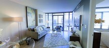 This contemporary single-floor residence showcases a bright, open-concept living area defined by expansive floor-to-ceiling windows that frame sweeping city views and flood the interior with natural light. The living room is thoughtfully appointed with hardwood flooring, a plush sectional sofa, and a glass-top coffee table, all anchored by a patterned area rug that adds warmth and texture to the space. A comfortable accent chair positioned near the windows creates an inviting reading nook bathed in natural light. The clean-lined, modern aesthetic is complemented by carefully curated wall art, adding color and personality to the neutral palette. A sleek media console anchors one side of the room, while a compact workspace with a glass desk tucks neatly against the windowed wall, making excellent use of the available space. The smooth, light-toned ceiling and hardwood floors lend the interior a polished, cohesive feel. The overall ambiance is bright, airy, and urban — a well-designed living space that balances comfort and sophistication, with the energy of the city as its backdrop.