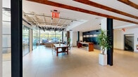 Located at 1409 W Broad St in Quakertown, PA, this single-floor space masterfully combines industrial character with contemporary design. The entrance hallway sets the tone immediately, featuring large-format light tile flooring, exposed wooden beam ceilings, and raw concrete walls that speak to the building's heritage. Glass double doors flood the entry with natural light, while a simple wooden bench provides a welcoming touch.

The expansive main area opens into a soaring, light-filled common space with an exposed steel ceiling structure and large floor-to-ceiling windows offering outdoor views. A built-in mailbox wall and a warm wood reception counter serve as functional focal points. Communal seating areas include a plush leather sofa grouping and a long communal dining table with modern chairs, fostering a collaborative atmosphere.

Steel-framed glass partitions divide portions of the space into semi-private zones, including a recreation room with hardwood floors and a shuffleboard table. These partitions maximize natural light while maintaining distinct areas.

The kitchen features sleek grey cabinetry, white quartz countertops, a stainless steel sink, and a striking dark subway tile backsplash, balancing practicality with refined aesthetics. Throughout, the interplay of exposed structural elements, modern furnishings, and abundant natural light creates a dynamic and inviting environment.