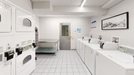 Located at 500 Glenelm Cres in Waterloo, Ontario, this shared laundry facility is a well-organized, single-room space designed for efficient, communal use. The room features white-tiled flooring and white walls that contribute to a clean, bright atmosphere, complemented by overhead fluorescent lighting that ensures clear visibility throughout.

The facility is equipped with a row of top-load washing machines, each outfitted with a Sparkle card-based payment terminal for convenient cashless transactions. Alongside the washers, stacked dryer units are positioned along the adjacent wall, maximizing the use of vertical space. A stainless steel utility sink is available for pre-treatment or hand-washing needs, and a sturdy stainless steel folding table provides a practical surface for sorting and folding laundry.

Visible ductwork and yellow-painted pipes run along the upper portion of the walls, reflecting the functional, utility-forward design of the space. Two grey access doors are present, one of which includes a window panel. Safety signage, including a "Safety First" notice and a wet floor caution sign, are prominently displayed. A framed piece of artwork on one wall adds a subtle decorative touch. Overall, the space is practical, well-maintained, and thoughtfully arranged to accommodate the laundry needs of residents.