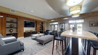 Located along Katy Freeway in Houston, Texas, this well-appointed clubhouse offers a thoughtfully designed single-floor layout that combines a fully equipped kitchen and an expansive living area. The open-concept floor plan flows seamlessly from one zone to the next, creating a welcoming and versatile communal environment.

The kitchen area is anchored by a large granite-topped island with dramatic veining, illuminated by a trio of cylindrical pendant lights. Dark espresso cabinetry lines the back wall, complemented by a tile backsplash and a mounted flat-screen TV, making it equally suited for casual gatherings or events.

The living area exudes a refined, contemporary aesthetic with a striking full-wall wood-paneled media unit featuring built-in shelving, dark cabinetry with stone countertops, and a centrally mounted flat-screen TV. Comfortable seating arrangements are anchored by patterned area rugs over light wood-look tile flooring, while large windows invite natural light and views of the surrounding landscape.

A secondary seating lounge provides additional intimate gathering space, adorned with framed artwork and access to the outdoors. Tray and coffered ceiling details, recessed lighting, and elegant drum pendant fixtures contribute to the polished, upscale ambiance throughout the entire space.