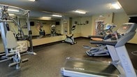 This single-level fitness room is designed to support a range of cardiovascular workouts in a functional, no-frills environment. The space is outfitted with Cybex-brand equipment, including treadmills, elliptical trainers, and a stationary bike, providing users with multiple cardio options. A large mirror spans one full wall, visually expanding the room while allowing users to monitor their form during exercise. The rubber flooring offers durability and comfort underfoot, making it well-suited for high-impact activity. Overhead fluorescent lighting ensures the room is evenly and brightly lit throughout. A wall-mounted television and a clock are also present, adding convenience for users during longer sessions. Natural light filters in through a window near the entrance door, lending a slightly more open feel to the interior. The overall layout is straightforward and practical, with equipment arranged to allow comfortable movement between machines. The neutral wall tones and utilitarian design keep the focus firmly on fitness functionality.