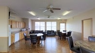 This single-floor community space offers a welcoming and functional environment designed for gathering and socializing. The open-concept layout is generously sized, providing ample room for multiple seating arrangements without feeling crowded. Several round dining tables, each surrounded by dark leather chairs, are positioned throughout the space, offering comfortable spots for meals or group activities. A dedicated lounge area features dark leather sofas and chairs arranged to encourage conversation and relaxation. The room is finished with warm hardwood-style flooring that adds a sense of cohesion and warmth throughout. Natural light pours in through a row of large windows along the exterior wall, brightening the neutral beige walls and creating an airy, open atmosphere. Overhead, ceiling fans with integrated lighting ensure consistent air circulation and comfort. Fluorescent light fixtures supplement the natural light, keeping the space well-lit throughout the day. A small kitchenette area is visible along one wall, equipped with cabinetry and a microwave, adding a practical convenience to the communal setting. An illuminated emergency exit sign is also present, reflecting attention to safety. Overall, this space is thoughtfully designed to serve as a versatile, comfortable community hub.