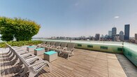 Located at 1282 Boylston Street in Boston's Fenway neighborhood, this rooftop deck offers an expansive open-air retreat with sweeping, unobstructed panoramic views of the Boston skyline, the Charles River, and the Cambridge waterfront. The deck is finished with warm wood planking throughout, lending a clean and contemporary aesthetic to the entire space.

The lounge area is anchored by a row of slatted Adirondack-style chairs paired with teal accent side tables, positioned directly along the glass perimeter railing to maximize the breathtaking cityscape views. The transparent glass balustrade ensures uninterrupted sightlines from every vantage point. Raised planters filled with lush flowering shrubs and leafy trees add vibrant greenery and a sense of privacy to the space.

A dedicated outdoor dining and grilling zone occupies one end of the deck, equipped with multiple gas grills, a mounted flat-screen television, and generous picnic-style dining tables with bench seating — ideal for social gatherings. Mature trees and tiered landscaped planters provide natural shade and a pleasant contrast against the building's modern paneled facade. Floor-to-ceiling glazed windows along the building wall reflect the open sky, further enhancing the airy, elevated atmosphere of this well-appointed rooftop amenity.