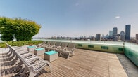 Located at 1282 Boylston Street in Boston's Fenway neighborhood, this rooftop deck offers an expansive open-air retreat with sweeping, unobstructed panoramic views of the Boston skyline, the Charles River, and the Cambridge waterfront. The deck is finished with warm wood planking throughout, lending a clean and contemporary aesthetic to the entire space.

The lounge area is anchored by a row of slatted Adirondack-style chairs paired with teal accent side tables, positioned directly along the glass perimeter railing to maximize the breathtaking cityscape views. The transparent glass balustrade ensures uninterrupted sightlines from every vantage point. Raised planters filled with lush flowering shrubs and leafy trees add vibrant greenery and a sense of privacy to the space.

A dedicated outdoor dining and grilling zone occupies one end of the deck, equipped with multiple gas grills, a mounted flat-screen television, and generous picnic-style dining tables with bench seating — ideal for social gatherings. Mature trees and tiered landscaped planters provide natural shade and a pleasant contrast against the building's modern paneled facade. Floor-to-ceiling glazed windows along the building wall reflect the open sky, further enhancing the airy, elevated atmosphere of this well-appointed rooftop amenity.