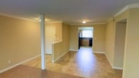 This single-floor residence offers a straightforward and functional layout designed for comfortable everyday living. The main living area is open and airy, featuring warm beige walls, clean white crown molding, and large-format stone-look tile flooring that extends throughout the space, lending a sense of continuity and cohesion. A structural support column with white trim adds a subtle architectural detail to the living area without interrupting the flow of the space. A half-wall partition with a pass-through opening separates the living zone from an adjacent area, enhancing the open-concept feel while providing a natural division between spaces. Flush-mount ceiling lighting provides consistent illumination across the main floor. Toward the rear of the residence, the kitchen features dark cabinetry with a traditional raised-panel design, a sink positioned beneath a window that allows natural light to filter in, and dark appliances including a refrigerator. The kitchen's layout offers practical workspace while maintaining visual connection to the rest of the home. White baseboards throughout complement the crown molding and contribute to a clean, classic interior aesthetic. Overall, the space presents a tidy, well-maintained environment with a neutral palette suited to a variety of furnishing styles.