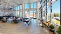 Located at 2000 South Blvd in Charlotte's South End neighborhood, this single-floor amenity lounge offers a thoughtfully designed communal space with distinct functional zones. The centerpiece of the space is a dramatic double-height great room, where soaring ceilings and floor-to-ceiling windows flood the interior with abundant natural light and frame views of the surrounding Charlotte skyline. A striking large-scale abstract mural anchors one wall, adding a bold artistic focal point. The lounge area features generous sectional seating arranged around low-profile wood coffee tables atop a teal area rug, complemented by high-top bistro tables and bar stools positioned near the expansive windows. A shuffleboard table adds a recreational element to the social space. Sculptural pendant chandeliers provide warm, layered lighting throughout. Adjacent to the lounge, a fully equipped kitchenette showcases navy-blue cabinetry with brass hardware, white countertops, a stainless steel sink, beverage refrigerator, microwave, and a tea and coffee station. The kitchenette's textured gray brick-patterned wall tile, warm wood-plank flooring, and brass wall sconces create an inviting, curated aesthetic consistent with the lounge's overall upscale, contemporary character.