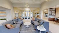 Located in Bradenton, Florida, this sophisticated single-story clubhouse serves as a premier community gathering space designed with contemporary coastal elegance. The welcoming entrance foyer features large, light-filled windows dressed with patterned drapery, herringbone-patterned tile flooring, and a comfortable bench seating area that sets the tone for the refined interiors within.

At the heart of the facility is a spacious lounge area framed by floor-to-ceiling glass partition walls, allowing natural light to flow freely throughout. Curved sectional sofas, accent chairs in teal and neutral tones, and statement pendant chandeliers create an inviting, upscale atmosphere. A separate seating area with small café tables and a built-in bench sits beneath a sweeping arched window, offering views of the outdoor amenity spaces.

The kitchenette is thoughtfully appointed with light wood cabinetry, quartz countertops, a wall-mounted flat-screen TV, a textured geometric tile backsplash, and stainless steel undercounter refrigerators—ideal for community events and gatherings. A beverage station with a coffee maker adds further convenience.

A dedicated game and entertainment lounge features a modern fireplace flanked by floating shelves, a wall-mounted TV, curated artwork, and a pool table, creating a relaxed yet sophisticated retreat. Private glass-enclosed meeting rooms offer intimate spaces for smaller gatherings, completing this well-appointed community facility.