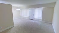 This single-floor space features an open living area that serves as the focal point upon entry. The room is finished with wall-to-wall neutral-toned carpeting, providing a soft and comfortable underfoot feel that extends throughout the space. A classic white paneled entry door is centrally positioned along the main wall, flanked on both sides by large windows dressed with vertical blinds that allow for adjustable natural light control. When fully open, these windows invite ample daylight into the room, enhancing the airy and open ambiance. The walls are finished in a soft, neutral tone that complements the light carpeting and contributes to the clean, understated aesthetic of the space. Overhead, an elegant chandelier with a traditional multi-arm design adds a refined decorative touch to the room, serving as a subtle focal point. White baseboards run along the perimeter, providing a crisp contrast to the carpeted floors. The overall design is straightforward and functional, offering a blank canvas that is well-suited for a variety of interior arrangements and décor styles.