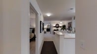 This single-floor residence is designed with an open and functional layout that seamlessly connects its key living spaces. A central hallway serves as the spine of the home, anchored by a dark patterned area rug that contrasts with the light-colored carpeted flooring running throughout. Crisp white walls and flush ceiling light fixtures create a clean, well-lit environment across the space.

To one side of the hallway, a bedroom is accessible through a wide doorway, featuring a plaid-upholstered bed and a glimpse of an ensuite or closet area beyond. Framed artwork lines the hallway wall, adding a decorative touch to the transitional space.

The hallway opens into a combined living and dining area, where warm ambient lighting from a pendant fixture contributes to an inviting atmosphere. A round decorative mirror and potted greenery add character to the living zone.

Adjacent to the living area, a kitchen features a wraparound breakfast bar with white countertops, providing both workspace and casual seating. The kitchen includes a sink and continues the home's consistent neutral palette, tying all areas together cohesively.