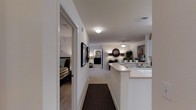 This single-floor residence is designed with an open and functional layout that seamlessly connects its key living spaces. A central hallway serves as the spine of the home, anchored by a dark patterned area rug that contrasts with the light-colored carpeted flooring running throughout. Crisp white walls and flush ceiling light fixtures create a clean, well-lit environment across the space.

To one side of the hallway, a bedroom is accessible through a wide doorway, featuring a plaid-upholstered bed and a glimpse of an ensuite or closet area beyond. Framed artwork lines the hallway wall, adding a decorative touch to the transitional space.

The hallway opens into a combined living and dining area, where warm ambient lighting from a pendant fixture contributes to an inviting atmosphere. A round decorative mirror and potted greenery add character to the living zone.

Adjacent to the living area, a kitchen features a wraparound breakfast bar with white countertops, providing both workspace and casual seating. The kitchen includes a sink and continues the home's consistent neutral palette, tying all areas together cohesively.