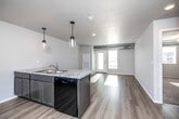 This well-appointed single-floor unit offers a thoughtfully designed open-concept layout that seamlessly connects the kitchen and living area. The kitchen is a standout feature, showcasing granite countertops, dark cabinetry, a peninsula island with a stainless steel sink and pendant lighting overhead, and a full suite of black appliances including a range, over-the-range microwave, and refrigerator. Light hardwood-style flooring flows throughout the main living areas, complementing the clean, neutral color palette and bright white walls. The living area is generously sized, bathed in natural light from glass panel doors that lead to the exterior. Exposed ductwork adds an industrial touch to the space. The bedrooms feature plush carpeting, windows for natural light, and ample wall space. The bathroom is clean and functional, with tile flooring, a dark-cabinet vanity with a granite top, and a full tub-shower combination. A dedicated laundry room with full-size washer and dryer and built-in shelving adds everyday convenience. Overall, the unit presents a modern, move-in-ready aesthetic with practical amenities and a cohesive design throughout.