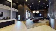 This upscale amenity lounge is defined by its dramatic double-height ceilings and a bold modern design palette of deep charcoal walls, warm wood-plank flooring, and expansive white marble-look tile. At the heart of the space stands a commanding cylindrical sculptural column clad in metallic mesh with a brass base, illuminated from within by a delicate chandelier — serving as a striking focal point upon entry. The main lounge area centers around a premium billiards table positioned beneath a pair of oversized globe pendant lights, creating an inviting atmosphere for social gatherings. Floor-to-ceiling glass doors open to an outdoor terrace, flooding the interior with natural light and providing seamless indoor-outdoor connectivity. A dedicated entertainment alcove features a large wall-mounted flat-screen TV surrounded by dark wood paneling, complemented by a full shuffleboard table and plush leather lounge chairs arranged for comfortable viewing. Bold pop-art wall pieces add a curated artistic touch to the space. A built-in bar area and additional seating niches with upholstered benches round out the lounge, making it a fully equipped, multi-functional social hub. The overall ambiance is refined yet energetic, blending luxury finishes with entertainment-forward design.
