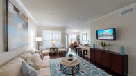 This single-floor residence offers a thoughtfully designed open-concept layout that seamlessly connects the main living areas. The living room serves as the central hub, featuring crown molding, rich dark hardwood flooring, and abundant natural light filtering through large windows with blinds. A teal-patterned area rug anchors the seating arrangement, while a wall-mounted flat-screen TV and a dark wood media console add functional style.

The dining area, separated by a breakfast bar with barstool seating, accommodates a glass-top dining table surrounded by woven rattan chairs beneath a track lighting fixture. Large windows allow ample natural light, complemented by lush greenery throughout.

The kitchen is well-equipped with dark cabinetry, granite-style countertops, and a full suite of stainless steel appliances, including a range, microwave, refrigerator, and dishwasher. An in-unit laundry area is conveniently accessible from the kitchen.

The bedroom features plush carpeting, a ceiling fan, a large dresser, and a window with natural views. A dedicated built-in workspace with a granite desktop, drawers, and overhead shelving provides an organized nook for productivity.

The bathroom offers a granite vanity, a large framed mirror, and a tiled shower-tub combination, completing this well-rounded residence.