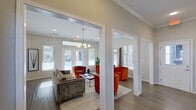This well-appointed single-floor community clubhouse and leasing office offers a thoughtfully designed layout that balances welcoming social spaces with functional professional areas. The open lounge area serves as the heart of the space, featuring rich hardwood-style flooring, crown molding, and an abundance of natural light streaming through large windows with white blinds. Bold orange velvet tufted seating arranged around sleek coffee tables creates a vibrant yet sophisticated atmosphere, while structural white columns add architectural character throughout. French doors open directly to an outdoor pool area, seamlessly connecting the interior to outdoor amenities. A dedicated lounge nook with a full-length mirror and additional seating provides a more intimate setting for conversation. The kitchenette and bar area is a standout feature, boasting dark cabinetry with granite countertops, pendant globe lighting, a wall-mounted TV, and a dramatic landscape mural that adds depth and visual interest. A carpeted conference room offers a quiet space for meetings, furnished with a long wooden table, modern white chairs, and views of the surrounding neighborhood. Private staff offices with carpeted floors, individual workstations, and framed artwork complete the professional side of the space. Throughout, crown molding and recessed lighting lend a polished, cohesive finish to the entire facility.