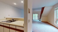 This two-floor residence at 1218 McKenna Blvd in Madison, Wisconsin offers a well-organized layout with a variety of functional living spaces across both levels. The main floor features a carpeted living area that flows openly toward a sliding glass door, providing access to an outdoor balcony and welcoming in natural light and greenery views. The kitchen is positioned adjacent to the living space with a pass-through counter design, white cabinetry with wood accents, tile flooring, and includes an electric range, refrigerator, and dishwasher. A full bathroom on the main level features a tub-shower combination, single vanity with storage, and tile flooring. The upper floor showcases dramatic vaulted ceilings that lend an open, airy feel to the space. A carpeted loft-style area overlooks the staircase with warm wood railing details. An upper-level room with soaring angled ceilings offers access to a private deck with open outdoor views of the surrounding Madison landscape. A ceiling fan is present on the upper level, complementing the wall-unit air conditioning. The staircase connecting both floors features classic wood balusters and serves as a central architectural element throughout the home.