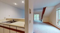 This two-floor residence at 1218 McKenna Blvd in Madison, Wisconsin offers a well-organized layout with a variety of functional living spaces across both levels. The main floor features a carpeted living area that flows openly toward a sliding glass door, providing access to an outdoor balcony and welcoming in natural light and greenery views. The kitchen is positioned adjacent to the living space with a pass-through counter design, white cabinetry with wood accents, tile flooring, and includes an electric range, refrigerator, and dishwasher. A full bathroom on the main level features a tub-shower combination, single vanity with storage, and tile flooring. The upper floor showcases dramatic vaulted ceilings that lend an open, airy feel to the space. A carpeted loft-style area overlooks the staircase with warm wood railing details. An upper-level room with soaring angled ceilings offers access to a private deck with open outdoor views of the surrounding Madison landscape. A ceiling fan is present on the upper level, complementing the wall-unit air conditioning. The staircase connecting both floors features classic wood balusters and serves as a central architectural element throughout the home.