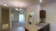 This single-floor residence offers a thoughtfully designed open-concept layout that connects the kitchen and living areas in a cohesive, functional flow. The kitchen is outfitted with rich dark wood cabinetry, a granite-look countertop, and a double stainless steel sink, providing ample prep and storage space. Tile flooring anchors the kitchen zone, transitioning smoothly into the carpeted living area, which features a ceiling fan and an elegant chandelier that adds a refined touch to the space. A sliding glass door in the living room allows natural light to fill the area and provides access to an outdoor space, enhancing the open and airy atmosphere. A dedicated in-unit laundry closet with a stacked washer and dryer adds everyday convenience and is neatly tucked between the kitchen and living zones. The neutral color palette throughout — from the warm beige carpet to the white walls and trim — creates a clean, welcoming ambiance. Recessed lighting and ceiling vents round out the practical yet polished character of this well-maintained single-level home.
