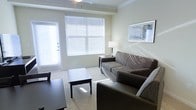 This single-floor space centers around a well-appointed living area designed for comfort and functionality. The room is characterized by large windows dressed with white horizontal blinds, allowing generous natural light to fill the space and creating a bright, airy feel. Crown molding along the upper walls adds a refined architectural detail, while a ceiling fan overhead provides practical climate control.

The living area is furnished with a combination of a fabric sofa and a dark leather accent chair, arranged around a rectangular dark-wood coffee table that anchors the seating arrangement. A matching end table beside the sofa holds a table lamp, offering warm ambient lighting. Neutral-toned carpeting runs throughout, complementing the soft beige wall color for a cohesive, understated palette. A framed piece of artwork adorns one wall, adding a subtle decorative touch.

A door with sidelight panel on the left side of the room provides access to an exterior area, further enhancing the sense of openness and connectivity to the outdoors. Overall, the space presents a clean, modern aesthetic with practical furnishings and a calm, neutral ambiance.