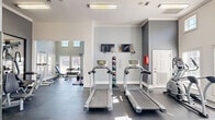 This fitness center in Columbia, SC, offers a comprehensive exercise facility designed to support a full range of workout routines. The space is housed within a single, open-plan exercise room featuring impressively high ceilings accented by elegant crown molding, lending the area a sense of openness and volume. Soft gray walls and dark flooring create a clean, modern aesthetic throughout.

Natural light floods the space through multiple large windows, including elevated clerestory windows that allow sunlight to reach deep into the room. Views of an outdoor pool area are visible from the free weights section, adding a pleasant outdoor connection. Wall-mounted flat-screen televisions are positioned near the cardio equipment for entertainment during workouts.

The cardio zone features multiple treadmills and an elliptical trainer, while a stationary bike and additional cardio machines occupy a separate section. The strength training area is well-appointed with a cable cross machine, a dumbbell rack with a full range of weights, a weight bench, and a barbell setup. A large wall mirror enhances the strength area, and a yoga mat and medicine ball rack provide options for functional fitness and stretching. Overhead fluorescent lighting ensures consistent illumination throughout the space.