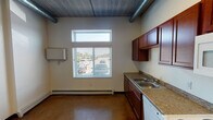Located at 1515 11th Ave N in Fargo, North Dakota, this single-floor unit blends industrial design elements with practical, everyday functionality. The space is characterized by its exposed ductwork and concrete ceilings, which lend a loft-like aesthetic throughout. Durable polished concrete flooring runs consistently across the unit, providing a sturdy and easy-to-maintain surface.

The kitchen is a standout feature, offering rich, warm-toned wood cabinetry extending from floor to ceiling for generous storage. Granite-patterned countertops provide ample workspace, complemented by a double-basin stainless steel sink, a full suite of white appliances including an electric range, over-the-range microwave, dishwasher, and refrigerator. A window with blinds allows natural light to brighten the kitchen area while offering views of the exterior.

A dedicated bedroom area includes a built-in closet with shelving and bi-fold doors, providing practical storage. The ceiling height in this area emphasizes the open, airy quality of the unit.

The bathroom is designed with accessibility in mind, featuring stainless steel grab bars positioned near the toilet and along the walls, a wide-clearance layout, a wood-toned vanity with an undermount sink, and a clean, minimalist finish throughout. Baseboard heating units are present throughout the unit, ensuring efficient climate control during Fargo's cold winters.