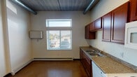 Located at 1515 11th Ave N in Fargo, North Dakota, this single-floor unit blends industrial design elements with practical, everyday functionality. The space is characterized by its exposed ductwork and concrete ceilings, which lend a loft-like aesthetic throughout. Durable polished concrete flooring runs consistently across the unit, providing a sturdy and easy-to-maintain surface.

The kitchen is a standout feature, offering rich, warm-toned wood cabinetry extending from floor to ceiling for generous storage. Granite-patterned countertops provide ample workspace, complemented by a double-basin stainless steel sink, a full suite of white appliances including an electric range, over-the-range microwave, dishwasher, and refrigerator. A window with blinds allows natural light to brighten the kitchen area while offering views of the exterior.

A dedicated bedroom area includes a built-in closet with shelving and bi-fold doors, providing practical storage. The ceiling height in this area emphasizes the open, airy quality of the unit.

The bathroom is designed with accessibility in mind, featuring stainless steel grab bars positioned near the toilet and along the walls, a wide-clearance layout, a wood-toned vanity with an undermount sink, and a clean, minimalist finish throughout. Baseboard heating units are present throughout the unit, ensuring efficient climate control during Fargo's cold winters.