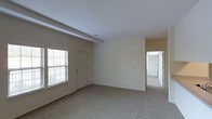 This single-floor space presents a straightforward, functional layout centered around a spacious main living area. The room is finished with wall-to-wall beige carpeting that adds a soft, understated quality to the interior. Neutral cream-colored walls and a white ceiling contribute to a bright, clean aesthetic throughout.

A window fitted with horizontal blinds allows natural light to filter into the living area, enhancing the open feel of the space. A white paneled door positioned near the window provides an exterior access point. Structural beam detailing is visible along the ceiling, adding a subtle architectural element to the otherwise minimalist design.

A visible HVAC vent in the ceiling indicates a central air system, ensuring climate control throughout. An electrical outlet is centrally positioned on the main wall, offering practical utility for the room.

An open doorway leads from the main living area into an adjoining room, which appears to continue the same neutral color palette and carpeted flooring. The connected layout suggests a seamless flow between spaces, making the overall floor plan feel cohesive and easy to navigate.