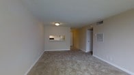 This single-floor space presents a modest yet functional layout centered around an open living area finished with wall-to-wall beige carpeting that lends a warm, understated feel underfoot. The room is defined by clean, neutral-toned walls and a textured popcorn ceiling, which adds a classic residential character. A flush-mount ceiling light fixture provides overhead illumination, while a wall-mounted HVAC vent ensures climate control convenience. A notable architectural feature is the built-in pass-through window connecting the living area to the adjacent kitchen, complete with a small shelf — a practical design element that promotes an open sightline between the two spaces. The kitchen beyond appears to feature its own lighting and cabinetry. A doorway visible toward the rear of the space leads to additional rooms, suggesting a straightforward and navigable floor plan. White baseboards frame the perimeter of the room, providing a clean finish against the carpeted floor. Overall, the space offers a simple, functional environment with practical features suited for everyday living.