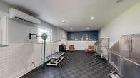 This purpose-built pet grooming studio is thoughtfully designed to accommodate professional grooming services in a clean and welcoming environment. The space is finished with white subway tile wainscoting along all walls, providing a hygienic and polished aesthetic that is both practical and visually appealing. Above the tile, light gray paint transitions to a bold teal accent wall adorned with playful, life-sized dog illustrations — adding warmth and personality to the professional setting.

The flooring throughout consists of interlocking dark checkered utility tiles, offering durability, slip resistance, and easy maintenance. Recessed lighting in the white ceiling ensures bright, even illumination across the entire workspace.

Two stainless steel elevated dog bathing tubs are positioned for efficient workflow, accompanied by a hydraulic lift grooming table complete with a grooming arm and restraints. A wall-mounted professional pet dryer adds to the suite of grooming equipment. A dark cabinetry unit with a butcher block countertop provides storage and a convenient workspace for tools and supplies. Two wooden slatted chairs offer a waiting area for clients. Natural light filters in through multiple windows, and a wall-mounted mini-split HVAC unit ensures year-round climate comfort for both pets and groomers.