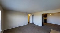 This single-floor residential unit offers a straightforward and functional layout designed for comfortable everyday living. The main living area is finished with wall-to-wall carpet flooring and neutral-toned walls, creating a clean and adaptable space that can accommodate a variety of furniture arrangements. A textured popcorn ceiling spans the room, and a ceiling-mounted HVAC vent provides climate control throughout the space.

From the living area, two doorways provide access to adjoining spaces. One leads to a closet, offering convenient built-in storage, while another opens to a warmly lit corridor or secondary room. On the opposite side of the living area, an open passage leads directly into the kitchen, which features dark wood cabinetry and what appears to be a refrigerator, providing a practical and efficient cooking space. The kitchen's darker tones contrast with the bright, light-filled living area, adding visual interest to the overall interior.

White baseboards frame the perimeter of the living space, contributing to a tidy and well-maintained appearance. The unit's open sightlines between the living area and kitchen enhance the sense of connectivity and ease of movement throughout the floor plan, making this a practical and livable single-floor space.