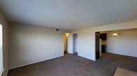 This single-floor residential unit offers a straightforward and functional layout designed for comfortable everyday living. The main living area is finished with wall-to-wall carpet flooring and neutral-toned walls, creating a clean and adaptable space that can accommodate a variety of furniture arrangements. A textured popcorn ceiling spans the room, and a ceiling-mounted HVAC vent provides climate control throughout the space.

From the living area, two doorways provide access to adjoining spaces. One leads to a closet, offering convenient built-in storage, while another opens to a warmly lit corridor or secondary room. On the opposite side of the living area, an open passage leads directly into the kitchen, which features dark wood cabinetry and what appears to be a refrigerator, providing a practical and efficient cooking space. The kitchen's darker tones contrast with the bright, light-filled living area, adding visual interest to the overall interior.

White baseboards frame the perimeter of the living space, contributing to a tidy and well-maintained appearance. The unit's open sightlines between the living area and kitchen enhance the sense of connectivity and ease of movement throughout the floor plan, making this a practical and livable single-floor space.