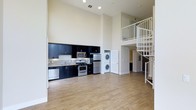 This thoughtfully designed two-story space makes a striking first impression with its soaring double-height ceilings that lend an airy, expansive feel to the main living area. The open-concept floor plan seamlessly connects the living space to a well-appointed kitchen, which features sleek dark cabinetry contrasted against light granite countertops, a tile backsplash, and a full suite of stainless steel appliances including a gas range, microwave, refrigerator, and dishwasher. Warm-toned hardwood flooring flows throughout the main level, adding a sense of continuity and natural warmth to the interior. A stacked washer and dryer unit is conveniently located in a dedicated nook on the main floor, offering practical in-unit laundry access. A standout architectural feature is the elegant white spiral staircase, which rises gracefully from the main level to the upper floor, where a mezzanine-style landing with white metal railings overlooks the living area below. The upper level provides additional living or sleeping space, accessible via the spiral stair. Recessed lighting illuminates the space efficiently, complementing the clean, modern aesthetic that defines the overall ambiance throughout both floors.